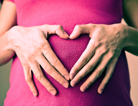 Close up of a pregnant woman in a pink top. Her hands make a heart over her baby bump