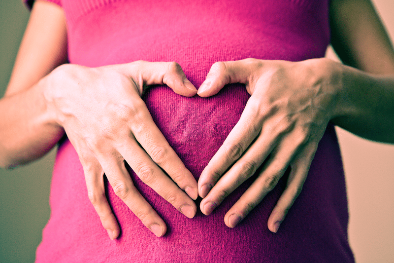 Close up of a pregnant woman in a pink top. Her hands make a heart over her baby bump