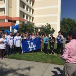 A group of Beverly Hospital employees stand together with their Dodgers flags and jerseys. A news station camera man in a pink shirt stands in the foreground.