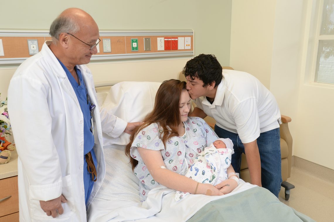 OBGYN Stephen Lee smiles as his patient holds her baby and receives a kiss on the forehead from her significant other