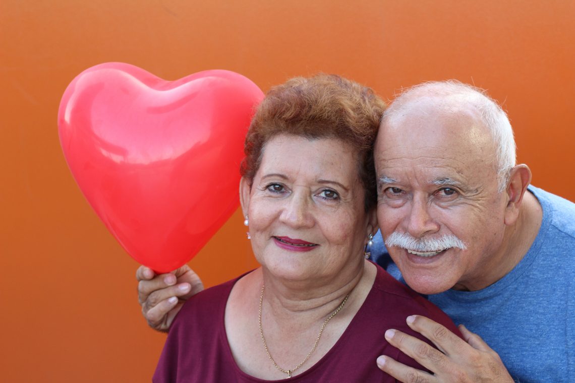 Senior hispanic couple smiling in front of orange background. Husband is holding a heart balloon .
