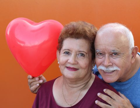 Senior hispanic couple smiling in front of orange background. Husband is holding a heart balloon .
