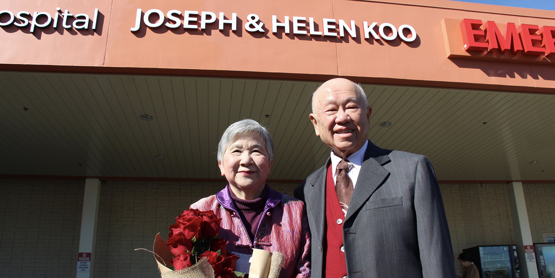 Mr. Joseph Loo and Dr. Helen Koo smile and pose under a sign with their names outside at the Emergency Room naming ceremony