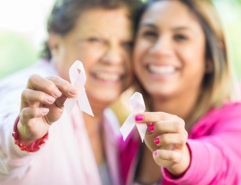 Senior Hispanic woman and adult daughter smile and hold up pink breast cancer awareness ribbons